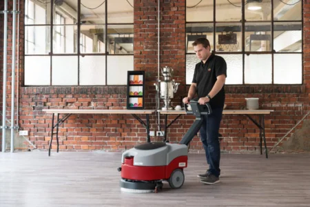 Mann reinigt einen Boden mit einer Scheuersaugmaschine in einem Raum mit Backsteinwand.